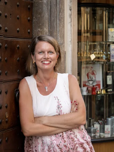 Bild von Simone Rieß, LinkedIn Mentoring und Ghostwriting. Zu sehen ist Simone, die lächelnd mit verschränkten Armen locker an eine rustikale braune Türe lehnt. Im Hintergrund ist ein Schaufenster mit Kafeezubehör zu sehen.